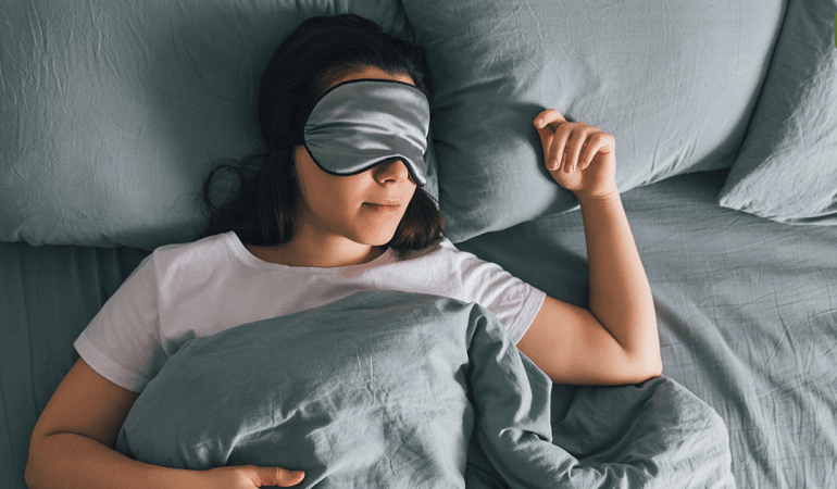 一名佩戴睡眠眼罩的女性在舒適的灰藍色床寢中熟睡，象徵透過環境控制提升 R.E.M. 快速動眼期質量。